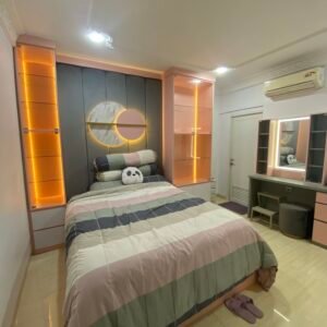 Interior kamar tidur dengan desain modern, tempat tidur berwarna pastel dengan pencahayaan LED oranye di kedua sisi dinding. Terdapat lukisan berbentuk lingkaran di dinding kepala tempat tidur, meja rias di sebelah kanan, dan lemari penyimpanan di sebelah kiri.