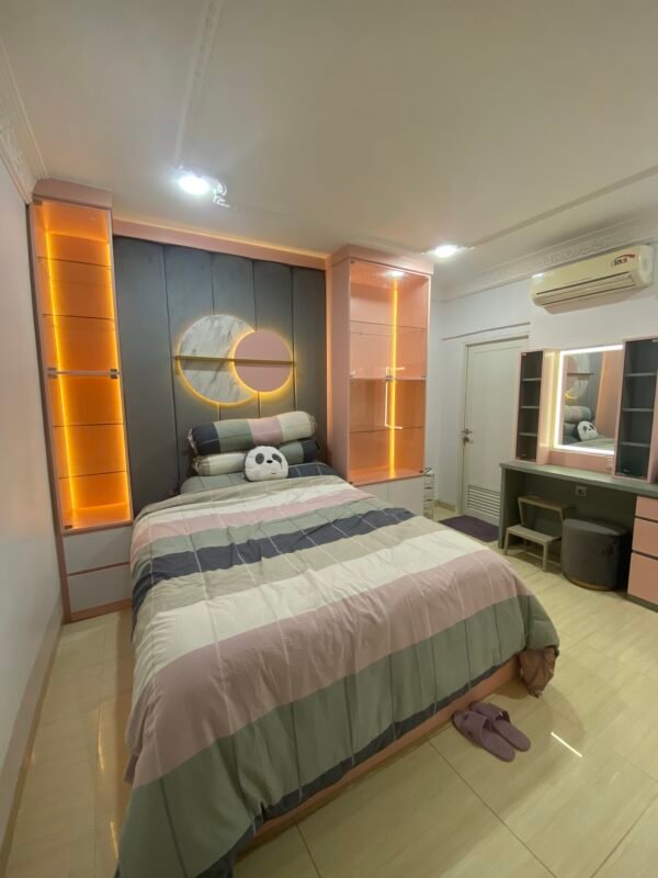 Interior kamar tidur dengan desain modern, tempat tidur berwarna pastel dengan pencahayaan LED oranye di kedua sisi dinding. Terdapat lukisan berbentuk lingkaran di dinding kepala tempat tidur, meja rias di sebelah kanan, dan lemari penyimpanan di sebelah kiri.